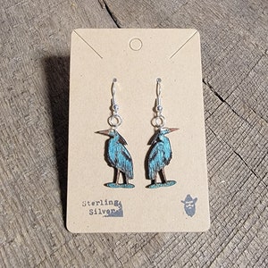 May include: A pair of sterling silver earrings featuring a blue heron design. The earrings are on a brown card with the text "Sterling Silver" printed on it.