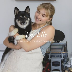 May include: A woman with blonde hair is holding a black and white dog in her arms. She is smiling and looking at the camera. The text "I'm Elle" and "Owner of Della Terra Goods & Design" is visible in the image.