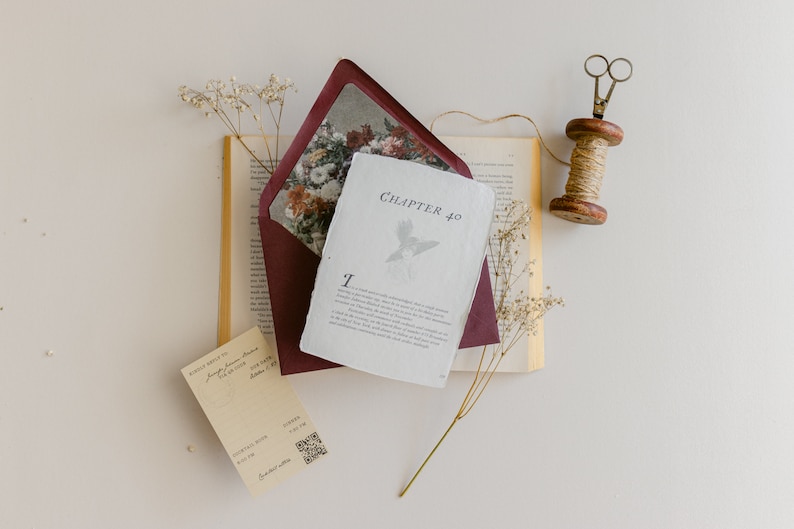 May include: A burgundy envelope with a floral design inside, a white card with the text "Chapter 40" and a bird illustration, a book with a white page, a spool of twine, and a pair of scissors on a white background.