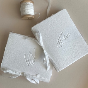 May include: Two white books with the words "Her Vows" and "His Vows" embossed on the covers. The books are tied together with a white ribbon.