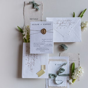 May include: Wedding invitation suite with a rustic, elegant theme. The invitation features a white card with black calligraphy, a wax seal, and a light blue ribbon. The envelope is made of textured paper and has a handwritten address. The text on the invitation reads "Aleah and Kaden" and "The wedding of Aleah and Kaden".