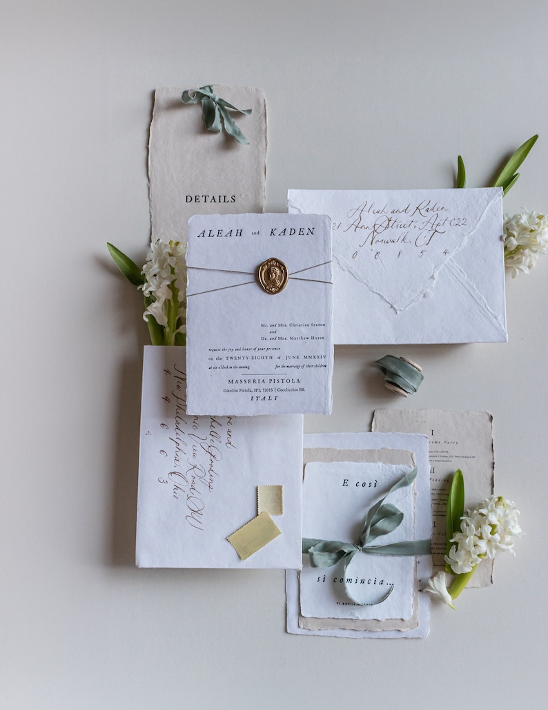 May include: Wedding invitation suite with a rustic, elegant theme. The invitation features a white card with black calligraphy, a wax seal, and a light blue ribbon. The envelope is made of textured paper and has a handwritten address. The text on the invitation reads "Aleah and Kaden" and "The wedding of Aleah and Kaden".
