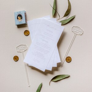 May include: A white menu card with a handwritten menu for a wedding reception. The menu includes a starter, entree, and dessert. The card is surrounded by eucalyptus leaves and gold wax seals.