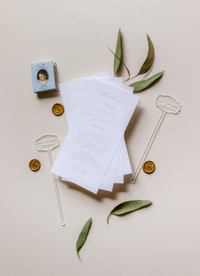 May include: A white menu card with a handwritten menu for a wedding reception. The menu includes a starter, entree, and dessert. The card is surrounded by eucalyptus leaves and gold wax seals.