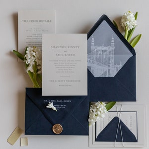 May include: A wedding invitation suite with a navy blue envelope, a white envelope with a gray inner liner, and a white card with the text "Shannon Kinney and Paul Rosen" and the details of their wedding at The Liberty Warehouse in Brooklyn, New York.