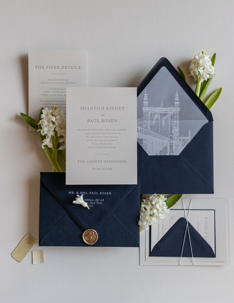 May include: A wedding invitation suite with a navy blue envelope, a white envelope with a gray inner liner, and a white card with the text "Shannon Kinney and Paul Rosen" and the details of their wedding at The Liberty Warehouse in Brooklyn, New York.
