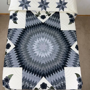 Puede incluir: Una colcha con un gran patrón de estrella central en tonos negro, gris y blanco. La colcha incluye motivos florales aplicados y una funda de almohada a juego con tres estrellas más pequeñas, todo sobre un fondo de color crema.
