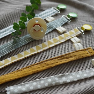 Könnte beinhalten: Fünf Stoff-Schnullerketten mit verschiedenen Mustern und Farben. Die Schnullerketten sind an Clips befestigt und haben kleine Etiketten mit dem Text "petit & tendre" darauf.