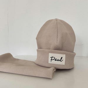 Könnte beinhalten: Beige geripptes Strickmützen- und Schal-Set. Die Mütze hat einen umgeschlagenen Rand mit einem weißen Etikett mit der Aufschrift "Paul" in schwarzer Schrift. Das Set ist aus einem weichen, neutralfarbenen Stoff gefertigt.