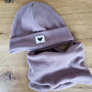 Könnte beinhalten: Ein Set aus lila Mütze und Halstuch. Die Mütze hat einen Umschlag und ein kleines weißes Quadrat mit einem schwarzen Herz. Beide Teile haben eine gerippte Struktur, was auf ein weiches Material hindeutet. Ideal für kalte Tage.