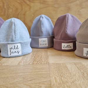 Könnte beinhalten: Fünf graue und lila gestrickte Mützen mit weißen Stoff-Etiketten. Die Etiketten tragen die Namen "Lina", "Wild Fang", "Ciao Kakao", "Lotte" und "Frida" in schwarzer Schrift.