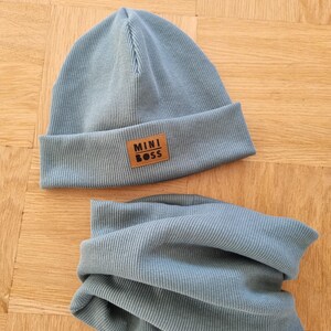 Könnte beinhalten: Ein zweiteiliges Set aus babyblauem Rippstrick, bestehend aus Mütze und Halstuch. Die Mütze hat einen umgeschlagenen Saum und einen kleinen braunen Lederaufnäher mit der Aufschrift "MINI BOSS". Das Halstuch ist röhrenförmig.