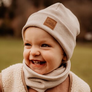 Könnte beinhalten: Ein hellbeige geripptes Strickmützen- und passendes Halstuch-Set. Die Mütze hat einen braunen Lederaufnäher mit dem Text "kleines Wunder". Das Kind trägt außerdem eine cremefarbene Sherpa-Weste und ein braunes Langarmshirt.