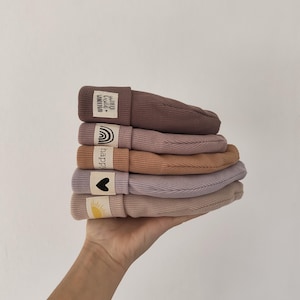 Könnte beinhalten: Ein Stapel von sechs gerippten Strickmützen in verschiedenen gedämpften Farben, darunter Mauve, Pfirsich und Beige. Jede Mütze hat ein kleines Stoffetikett mit einer Grafik oder Text, wie einem Regenbogen, Herz oder Sonne. Die Mützen werden in einer Hand vor weißem Hintergrund gehalten.