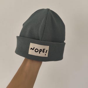 Könnte beinhalten: Eine tealfarbene Mütze aus Rippstrick mit umgeschlagenem Saum. Ein rechteckiger Aufnäher mit dem Wort "NOPE!" in schwarzer Schrift ist auf der Vorderseite aufgenäht. Die Mütze ist aus weichem, dehnbarem Material.