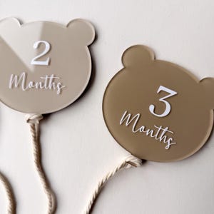 May include: Two bear-shaped monthly milestone markers. One marker is light beige with the number "2" and the word "Months" in white script. The other is brown with the number "3" and the word "Months" in white script. Each has a cream-colored string.