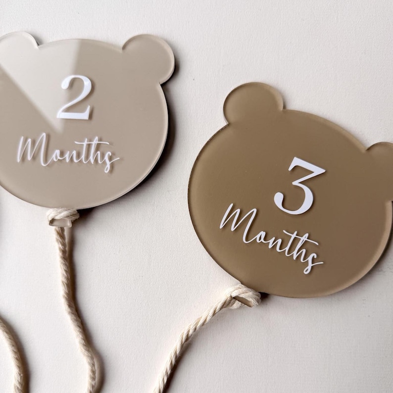 May include: Two bear-shaped monthly milestone markers. One marker is light beige with the number "2" and the word "Months" in white script. The other is brown with the number "3" and the word "Months" in white script. Each has a cream-colored string.
