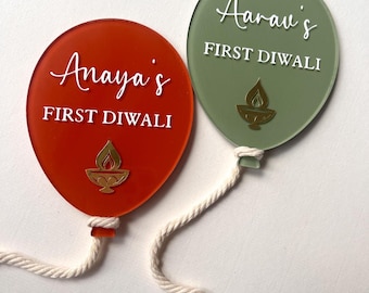 May include: Two balloon-shaped acrylic ornaments in orange and sage green. The orange ornament features the white text Anaya's FIRST DIWALI and a gold diya lamp. The green ornament features the white text Aarav's FIRST DIWALI and a gold diya lamp.