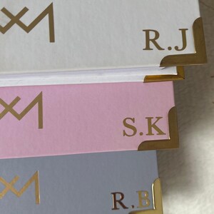 May include: Three notebooks with gold accents and initials. The top notebook is white with the initials "R.J." The middle notebook is pink with the initials "S.K." The bottom notebook is light blue with the initials "R.B."