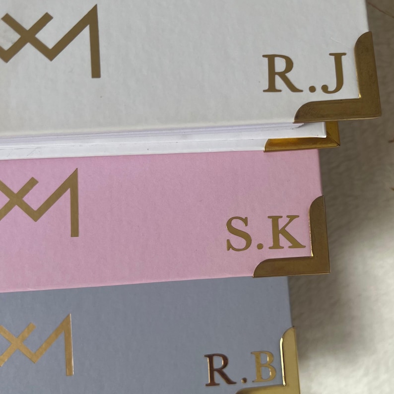 May include: Three notebooks with gold accents and initials. The top notebook is white with the initials "R.J." The middle notebook is pink with the initials "S.K." The bottom notebook is light blue with the initials "R.B."