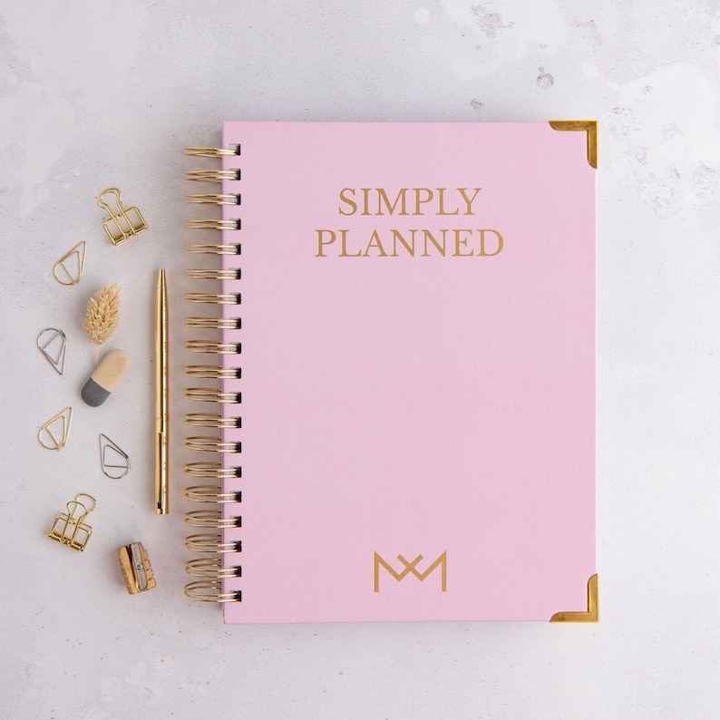 May include: A pink spiral bound planner with gold accents and the text "SIMPLY PLANNED" in gold lettering. The planner has a gold monogram of "M&M" on the bottom right corner.