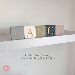May include: Five square wooden blocks arranged in a row spelling the name VANCE. Each block features a different color: snowflake blue, soft ivory, gentle olive, deep blue, and soft grey. The letters are engraved in a metallic copper finish.