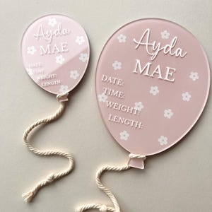 May include: Two pink acrylic balloon-shaped baby announcement signs with white floral designs and the name "Ayda MAE" written in white cursive. Each sign has a white rope attached to the bottom.