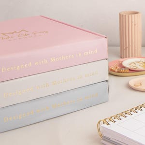 May include: Three stackable boxes in a light pink, white and light blue colour scheme. The boxes are labelled with gold lettering that reads "Designed with Mothers in mind".