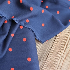 K&ouml;nnte beinhalten: Marineblauer Stoff mit einem Muster aus leuchtend roten Polka-Dots. Der Stoff ist drapiert und zeigt eine rohe Kante. Er liegt auf einer hellbraunen Holzoberfl&auml;che. Der Stoff hat ein rotes Druckdesign.