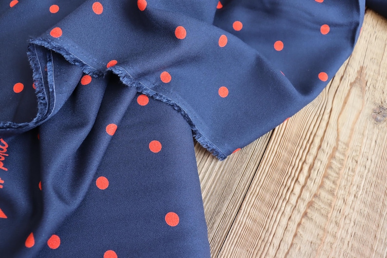 K&ouml;nnte beinhalten: Marineblauer Stoff mit einem Muster aus leuchtend roten Polka-Dots. Der Stoff ist drapiert und zeigt eine rohe Kante. Er liegt auf einer hellbraunen Holzoberfl&auml;che. Der Stoff hat ein rotes Druckdesign.