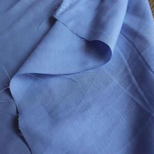 May include: A close-up of a light blue fabric with a subtle sheen. The fabric is folded over, revealing its texture and the way it drapes. The edges show the raw, unfinished cut of the material. The background is a wooden surface.