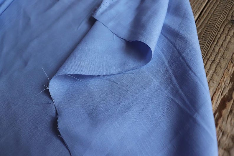 May include: A close-up of a light blue fabric with a subtle sheen. The fabric is folded over, revealing its texture and the way it drapes. The edges show the raw, unfinished cut of the material. The background is a wooden surface.
