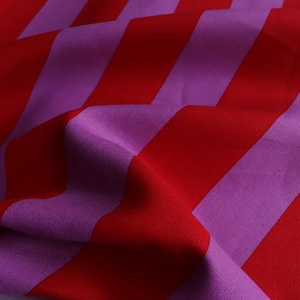 Può includere: Primo piano di un tessuto con un audace motivo a righe diagonali. Il tessuto presenta strisce alternate di rosso vibrante e viola tenue. Il materiale appare morbido e fluido, con pieghe e pieghe visibili.