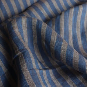 May include: Close-up of a blue and gray striped fabric. The fabric is draped, showing the texture and pattern of the material. The stripes are vertical and evenly spaced, creating a classic design.