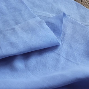 May include: Close-up of a light blue fabric with a subtle sheen. The fabric is folded, creating soft shadows and highlighting the texture. The material appears lightweight and possibly linen or silk.