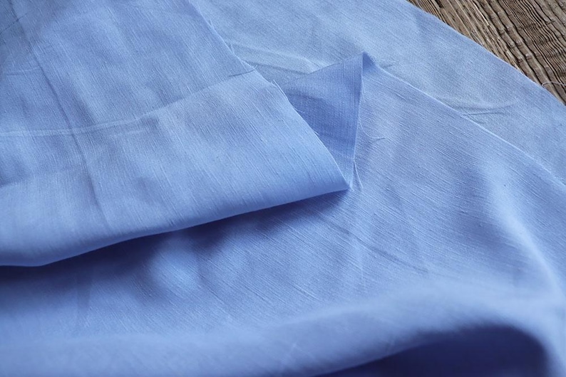 May include: Close-up of a light blue fabric with a subtle sheen. The fabric is folded, creating soft shadows and highlighting the texture. The material appears lightweight and possibly linen or silk.