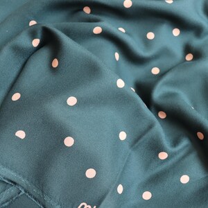 Puede incluir: Tela color turquesa con un patr&oacute;n repetitivo de lunares rosa claro. La tela est&aacute; doblada, creando sombras y textura. El material parece suave y fluido, adecuado para bufandas o prendas.