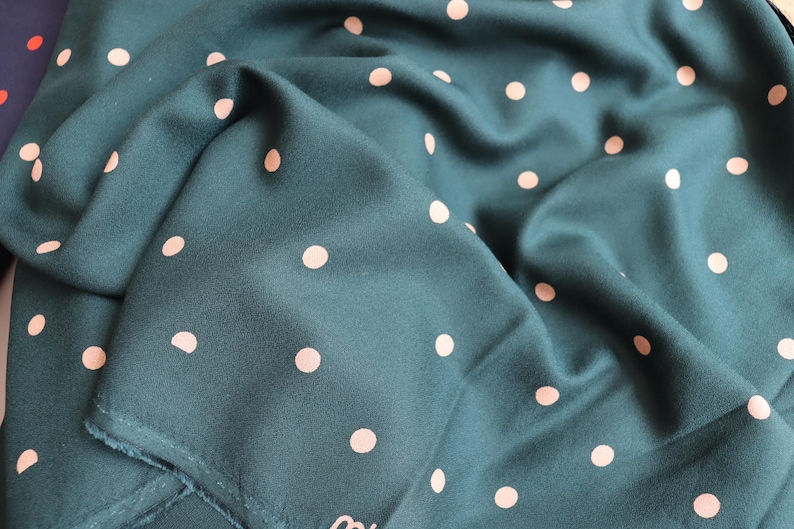 Puede incluir: Tela color turquesa con un patr&oacute;n repetitivo de lunares rosa claro. La tela est&aacute; doblada, creando sombras y textura. El material parece suave y fluido, adecuado para bufandas o prendas.