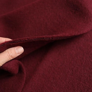 Könnte beinhalten: Nahaufnahme einer Hand, die einen Abschnitt aus bordeauxfarbenem Stoff hält. Der Stoff hat ein weiches, strukturiertes Aussehen und eine sichtbare Naht am Rand. Die Farbe ist ein tiefer, satter Rotton.