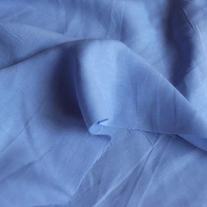 May include: Close-up of a light blue fabric with soft folds and a slightly textured surface. The fabric is a solid color, with a subtle sheen, and appears to be a lightweight material, possibly linen or cotton.