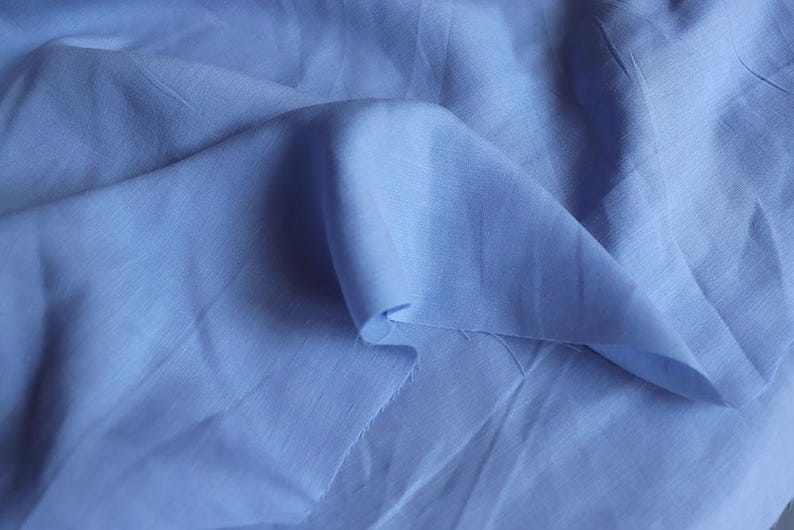 May include: Close-up of a light blue fabric with soft folds and a slightly textured surface. The fabric is a solid color, with a subtle sheen, and appears to be a lightweight material, possibly linen or cotton.