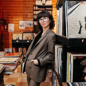 Op de afbeelding: Een persoon in een bruin geruit jasje en bijpassende broek, leunend tegen een plank met vinylplaten. De setting is een vintage platenzaak met houten lambrisering en planken vol albums. De persoon draagt een bril en een zwart shirt.