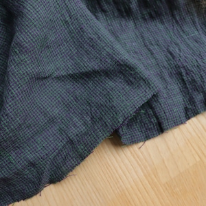 Könnte beinhalten: Nahaufnahme eines dunkelgrauen Stoffes mit einem kleinen Karomuster in Grün und Lila. Der Stoff ist leicht zerknittert und hat einen rohen Rand. Das Material scheint Leinen oder ein ähnliches Webmaterial zu sein.