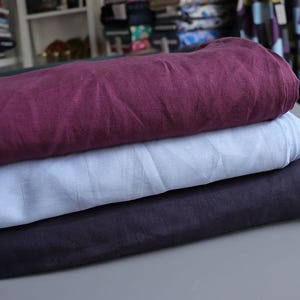 May include: A stack of three folded fabric pieces in shades of burgundy, light blue, and dark navy. The fabrics appear to be linen or a similar natural fiber, with a slightly wrinkled texture. The fabrics are stacked on a gray surface.