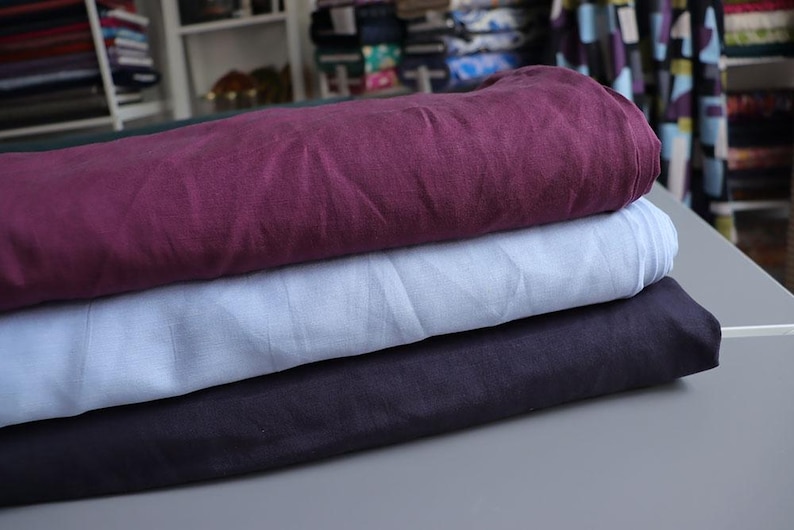 May include: A stack of three folded fabric pieces in shades of burgundy, light blue, and dark navy. The fabrics appear to be linen or a similar natural fiber, with a slightly wrinkled texture. The fabrics are stacked on a gray surface.