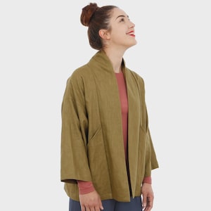 May include: Olive green kimono-style jacket with wide sleeves and a relaxed fit. The jacket has a shawl collar and two front pockets. The model is wearing a red long-sleeve shirt underneath. The jacket is made of a textured fabric.