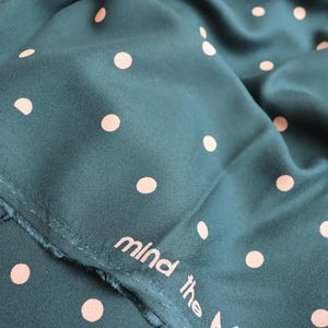 Puede incluir: Primer plano de una tela verde azulado con un patr&oacute;n repetido de lunares color melocot&oacute;n. La tela est&aacute; doblada, mostrando la textura y un borde sin rematar. Las palabras "mind the" est&aacute;n impresas en la tela.