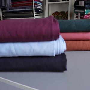 May include: A stack of folded fabric rolls in various colors, including deep purple, light blue, and dark navy. Other colors include coral, rust, and teal. The fabrics are displayed on a gray surface, with shelves of more fabric in the background.