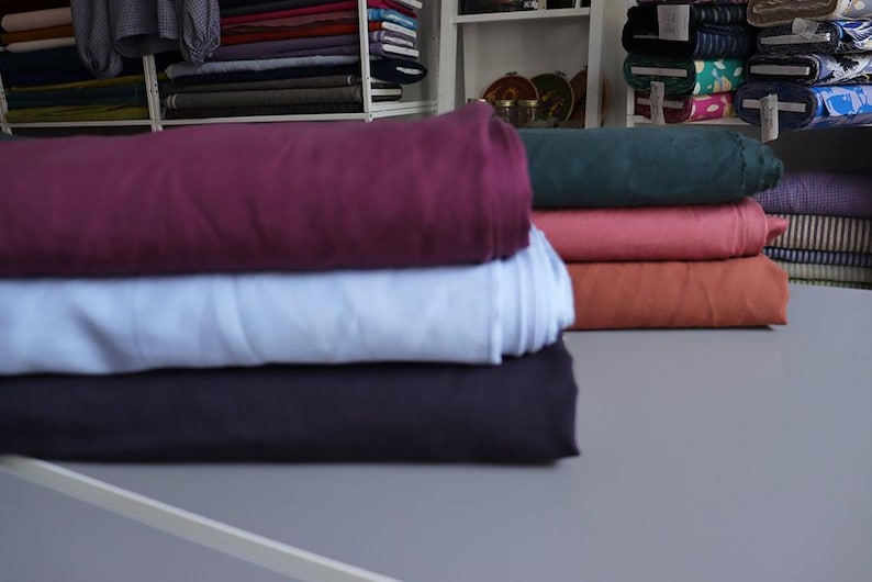 May include: A stack of folded fabric rolls in various colors, including deep purple, light blue, and dark navy. Other colors include coral, rust, and teal. The fabrics are displayed on a gray surface, with shelves of more fabric in the background.