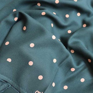 Puede incluir: Una tela de color verde azulado con un patr&oacute;n repetido de lunares rosa claro. La tela est&aacute; doblada y drapeada, mostrando su textura y la forma en que captura la luz. El efecto general es suave y fluido.